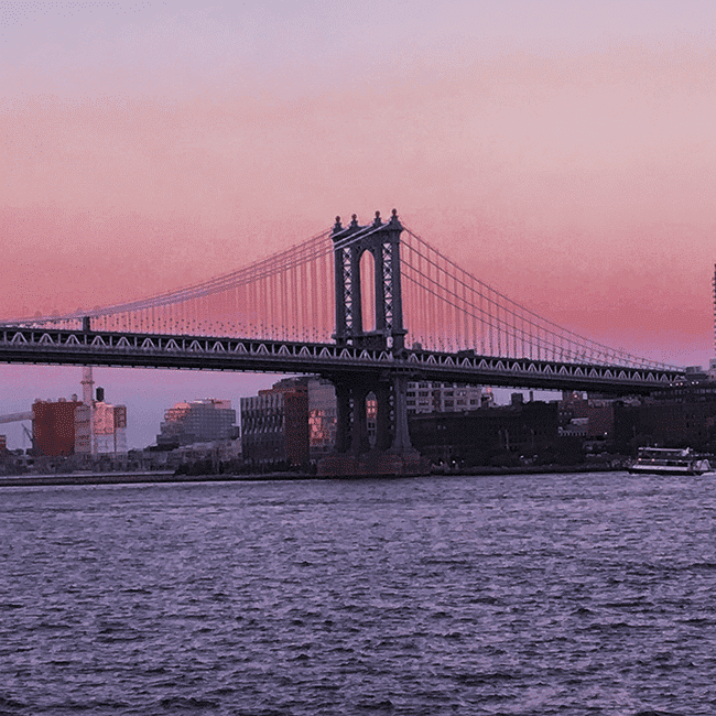 Image shows the Brooklyn Bridge at dusk. The sky is pink and purple, and the water of the East River is a deep, dark blue.