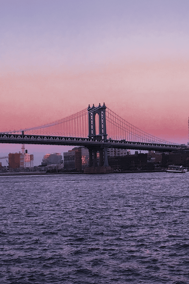 Image shows the Brooklyn Bridge at dusk. The sky is pink and purple, and the water of the East River is a deep, dark blue.
