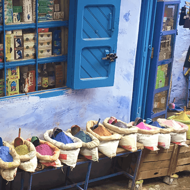The image shows the window of a Moroccan drugstore. The medina and windows are painted blue, and brightly coloured pigments are visible below the window.