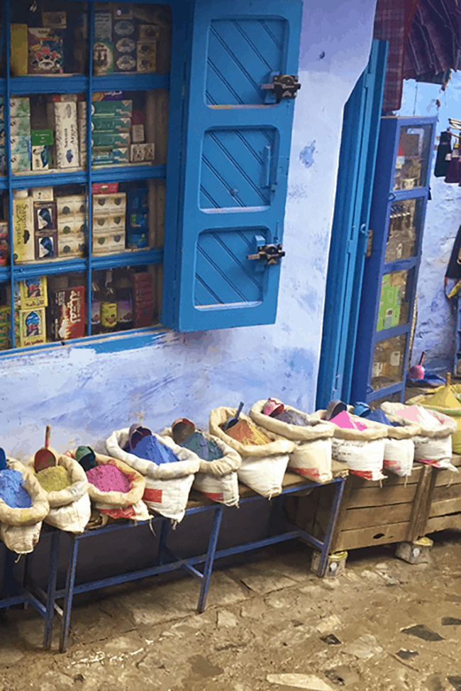 The image shows the window of a Moroccan drugstore. The medina and windows are painted blue, and brightly coloured pigments are visible below the window.