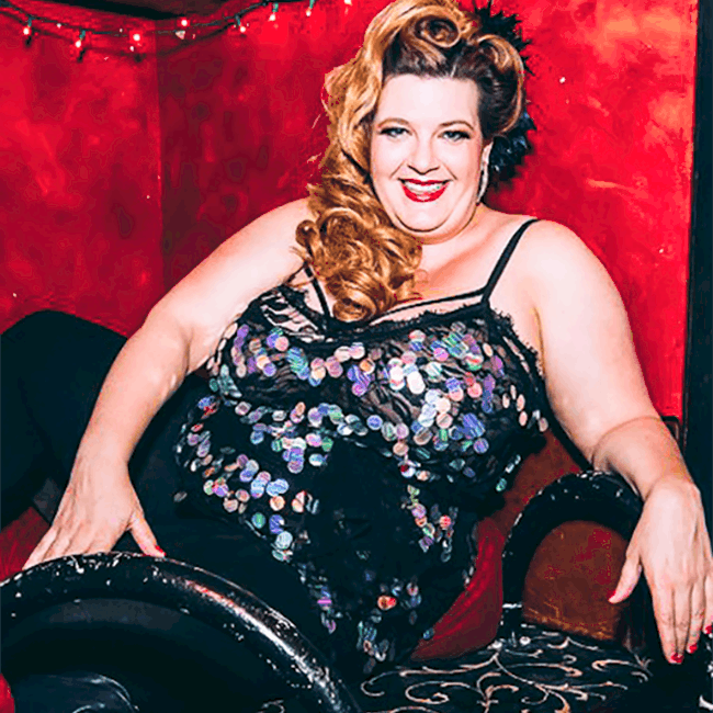 Image shows Jezebel sitting in a velvet cubbyhole at a New York city bar, smiling and wearing a short black dress covered in sparkly silver pailettes.