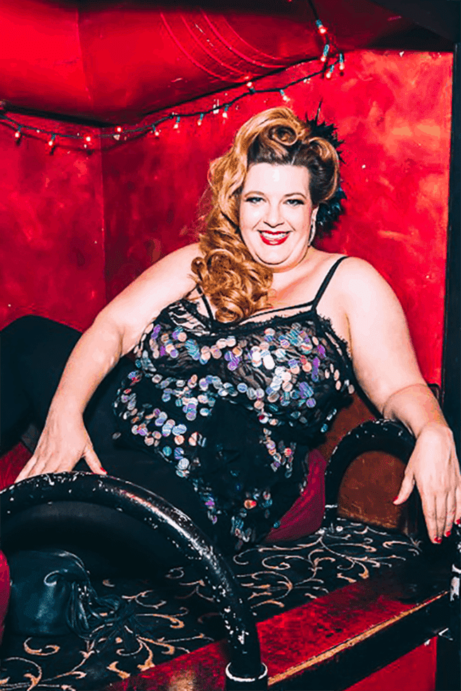 Image shows Jezebel sitting in a velvet cubbyhole at a New York city bar, smiling and wearing a short black dress covered in sparkly silver pailettes.