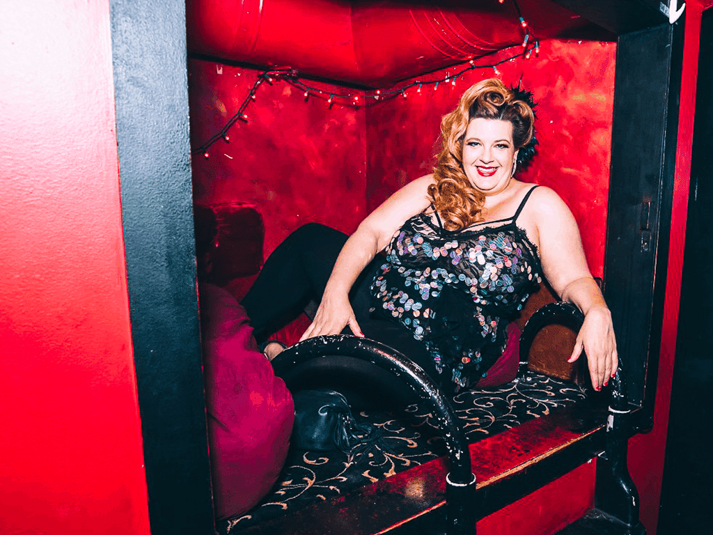 Jezebel sits in a velvet cubbyhole at a nighclub, wearing a fancy dress. She is smiling out at the viewer conversationally.