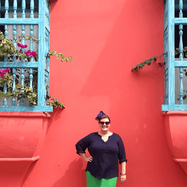 Image shows Jezebel standing in front of a pink building smiling. She is wearing a navy blue top and a bright green skirt which the wind is blowing. The photo exists above the copy "How To Make Travel Easier For People of Size"