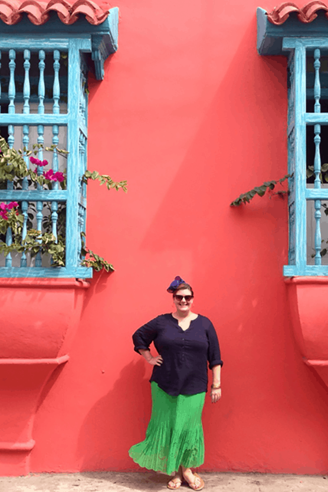 Image shows Jezebel standing in front of a pink building smiling. She is wearing a navy blue top and a bright green skirt which the wind is blowing. The photo exists above the copy "How To Make Travel Easier For People of Size"