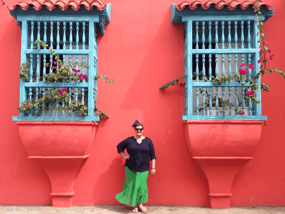 Image shows Jezebel standing in front of a pink building smiling. She is wearing a navy blue top and a bright green skirt which the wind is blowing. The photo exists above the copy "How To Be An Ally To Plus Size Travelers"
