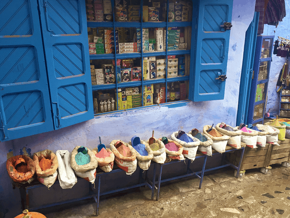 Image shows the medina in the blue city of Chefchaouen. Multiple colours of pigment are arranged against a blue wall. Image appears above article outlining cheap travel essentials.