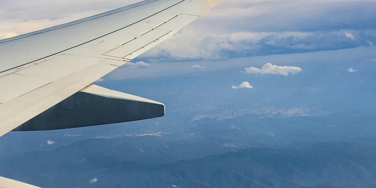 Image shows the view of liftoff from an airlplane window seat. Clouds are visible above craggy green mountains.