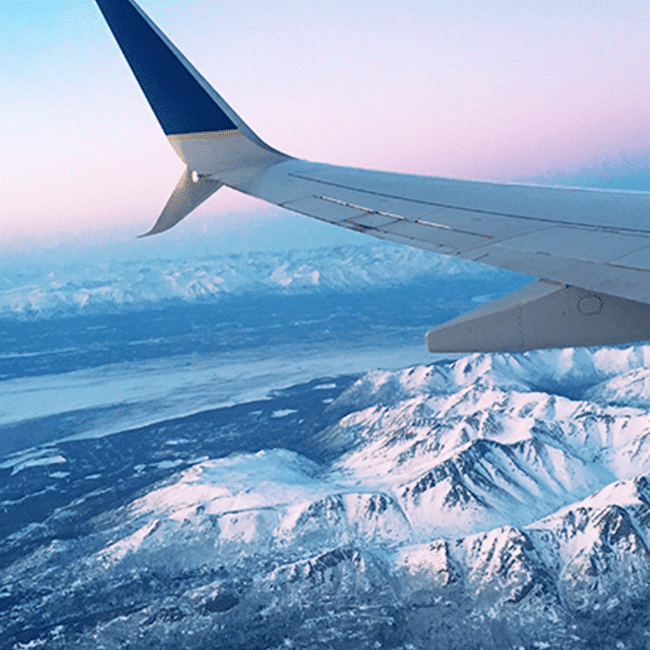 Image shows the wing of a plane with a view of snow-sovered mountains below it.