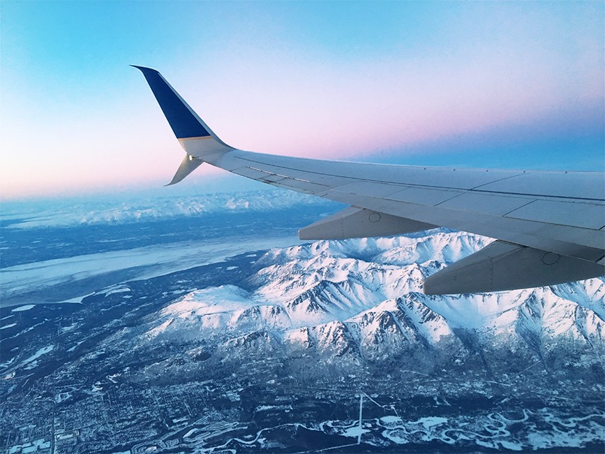 Image shows the wing of a plane with a view of snow-sovered mountains below it.