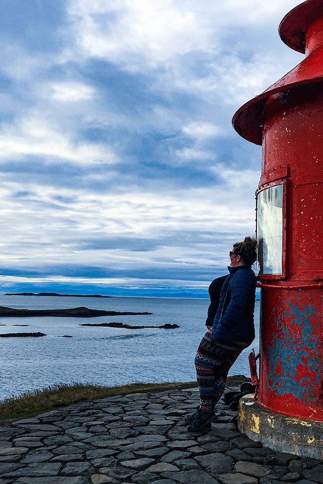 Jezebel is standing against a red lighthouse, looking out at the ocean. She is wearing hiking gear and a blue coat. The sky is scattered with clouds.