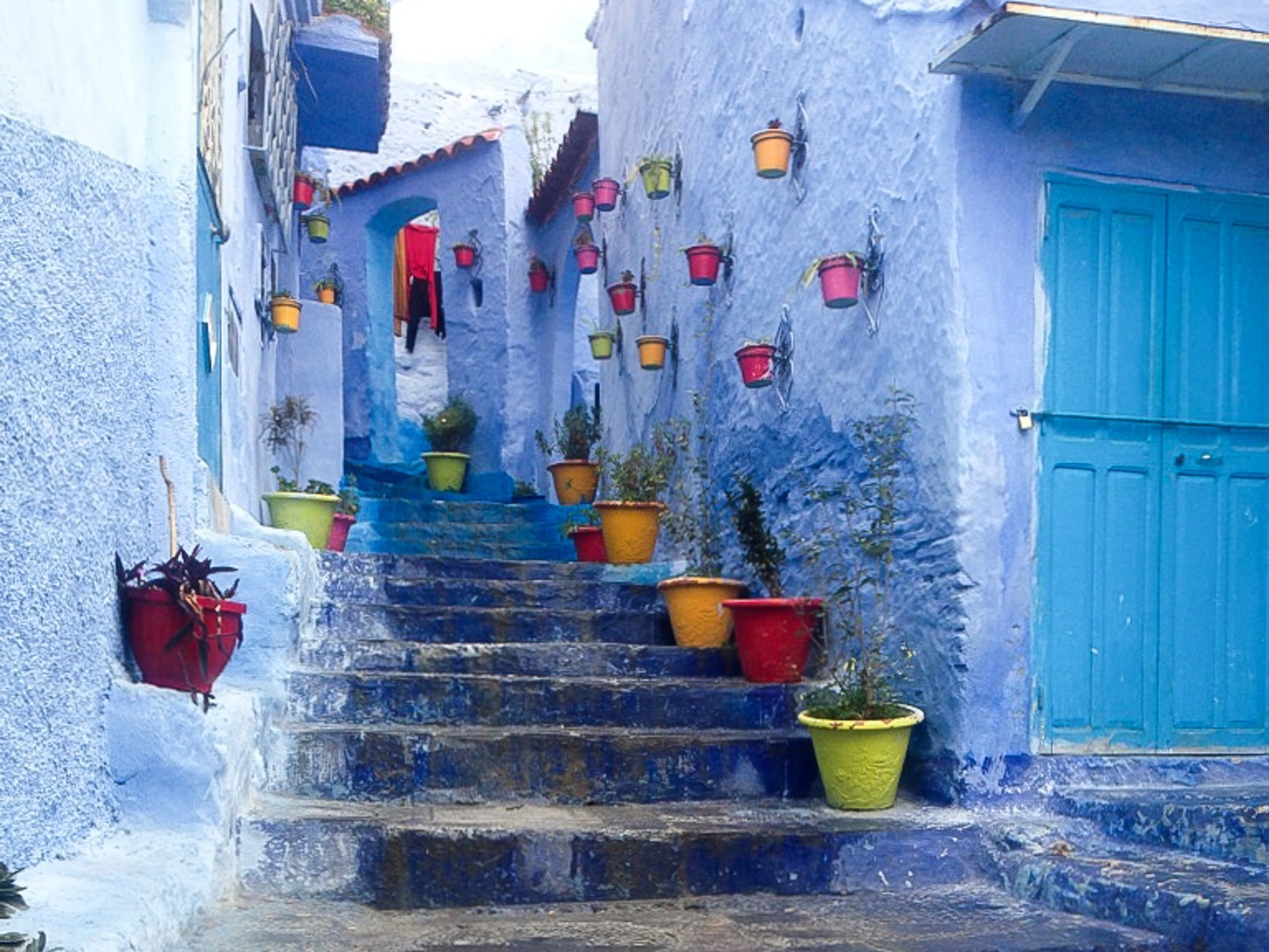 Image shows a stairwell in the Moroccan city of Chefchaouen. The medina is painted blue, with doors and stairs painted different shaes of blue. Along the stairwell, brightly coloured flowerpots hang against walls.